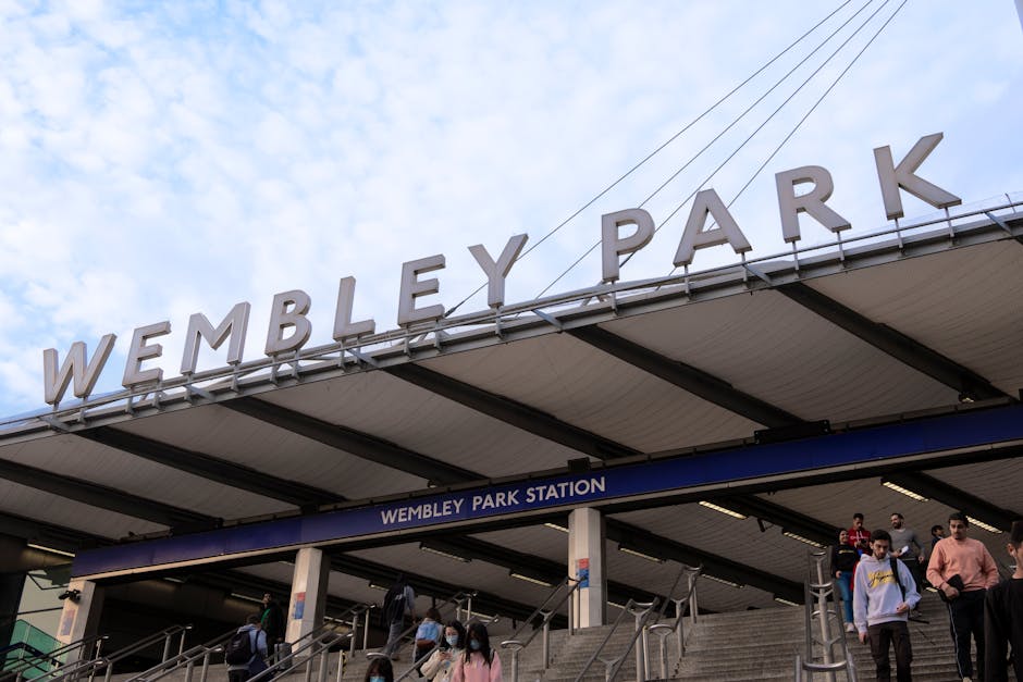 The image shows the exterior of Wembley Park station with a large sign displaying 'WEMBLEY PARK' in bold, white uppercase letters mounted on the station's roof. Below the sign, there is a blue sign indicating 'WEMBLEY PARK STATION' attached to the front of the entrance canopy. The station entrance features a set of stairs with metal handrails on both sides, and several people are seen ascending and descending the stairs, some wearing face masks. The sky above is partly cloudy with patches of blue visible. The station setting and the presence of pedestrians illustrate a busy urban transit environment, relevant to house removals and moving logistics.