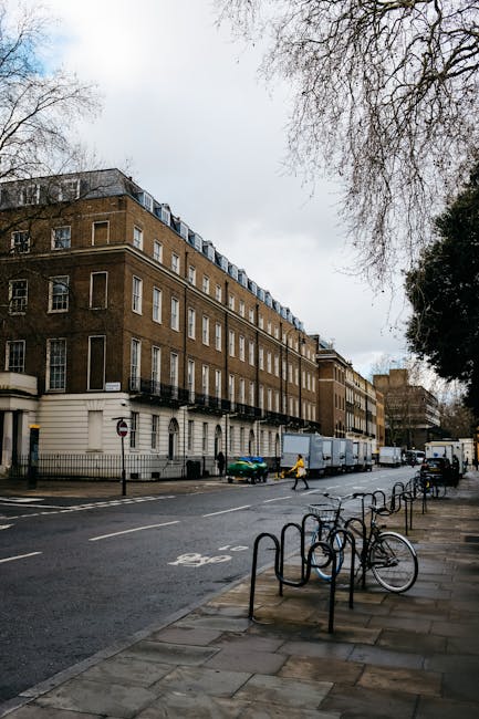 A street view of an urban residential area with a large, multi-storey brick building featuring white window frames and a black iron balcony on the first floor. In the foreground, there are several met